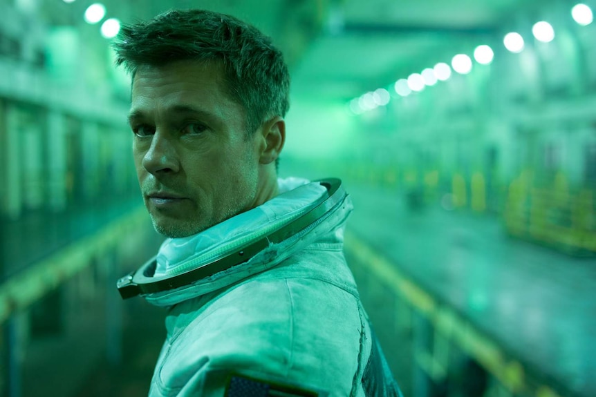 Close-up of Brad Pitt in green-lit space-type hold, wearing space suit but no helmet, looking at camera.