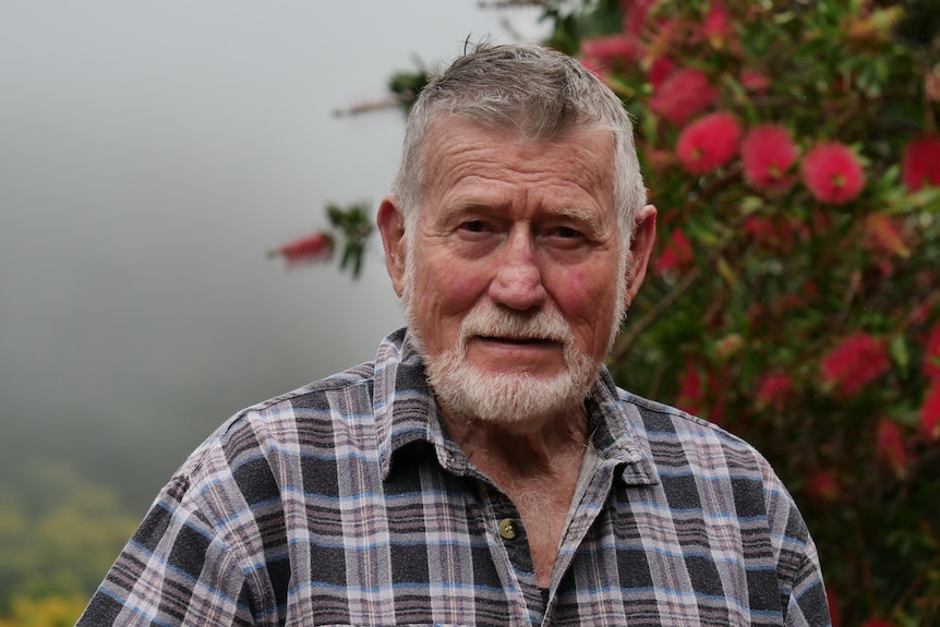 A man with grey hair and a checked shirt stands in front of trees and mist on a farm.