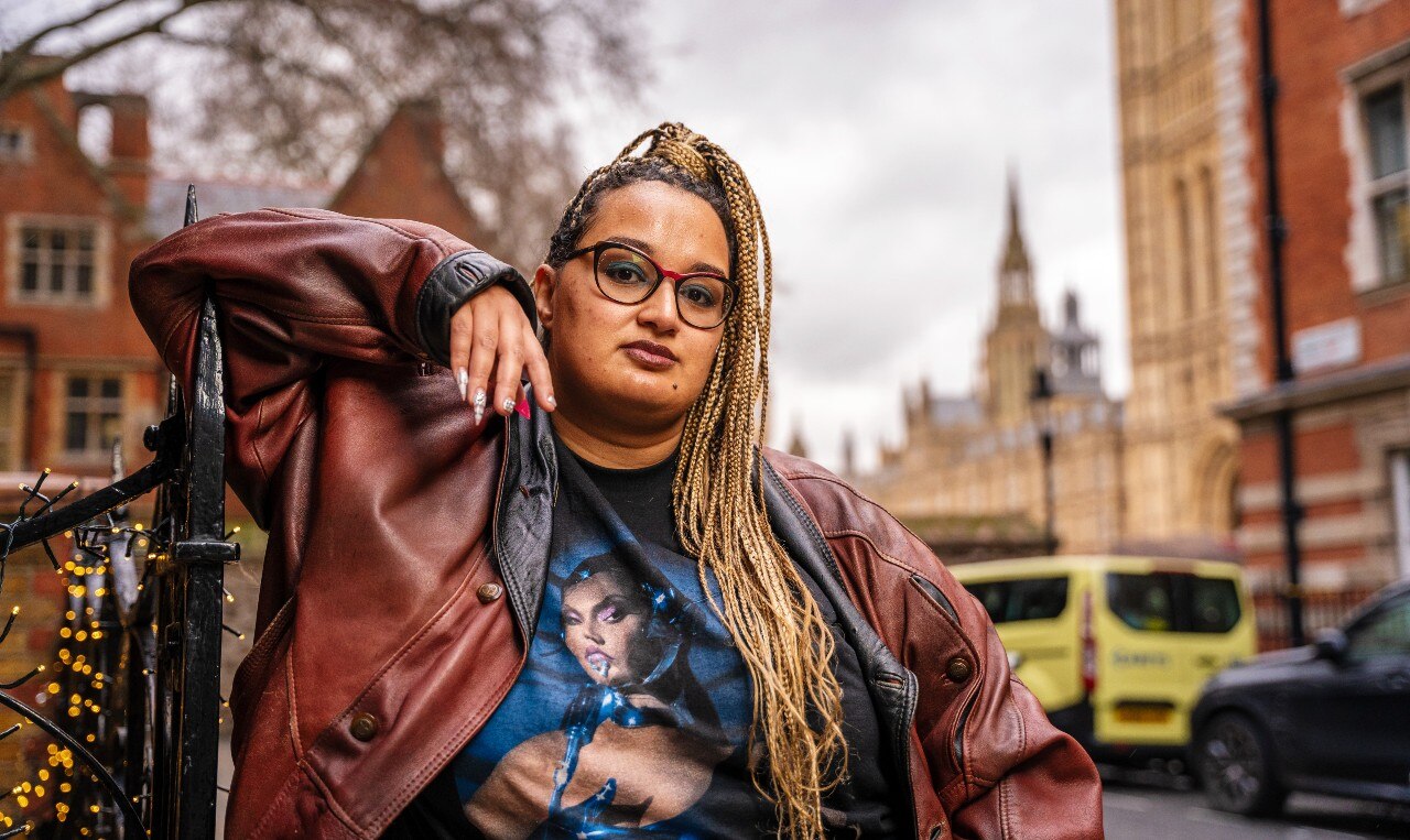 A woman with a neutral expression leans on a fence and looks at the camera. She is surrounded by historic buildings