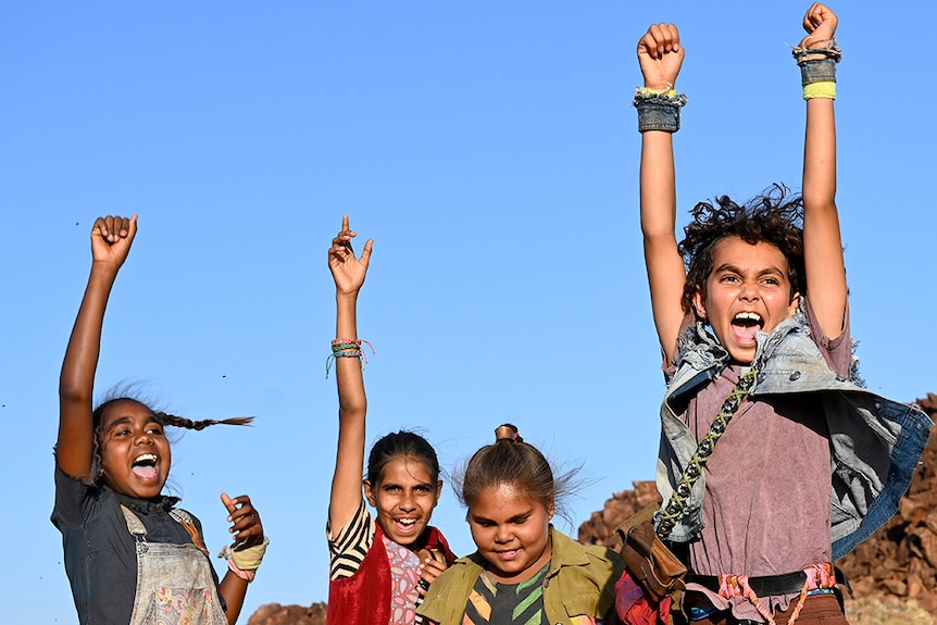 Six children raise hands and fist in the air and celebrate, one boy is jumping, in rocky desert landscape with spinifex.