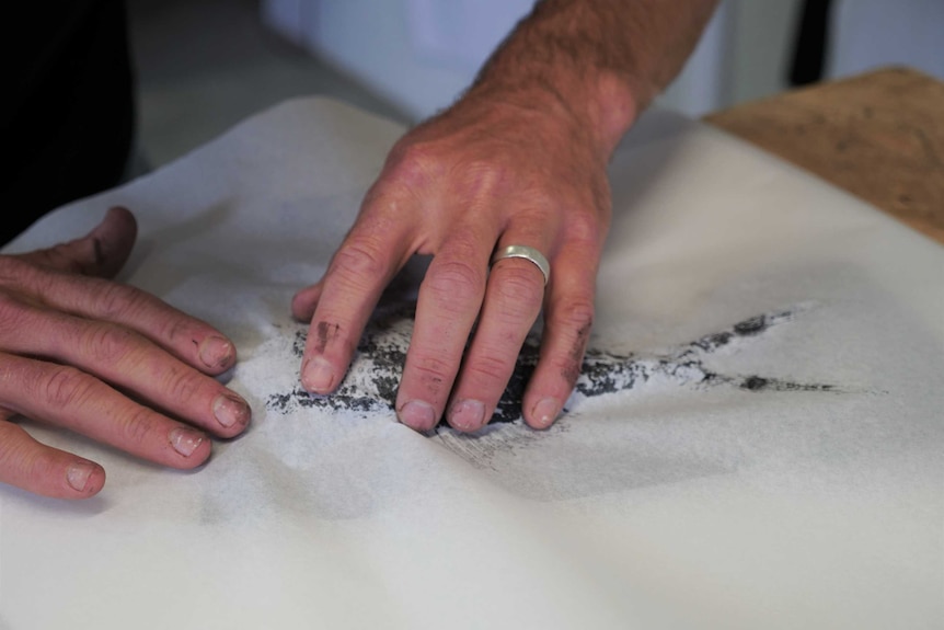 A pair of hands press a piece of paper onto an inked dead fish.