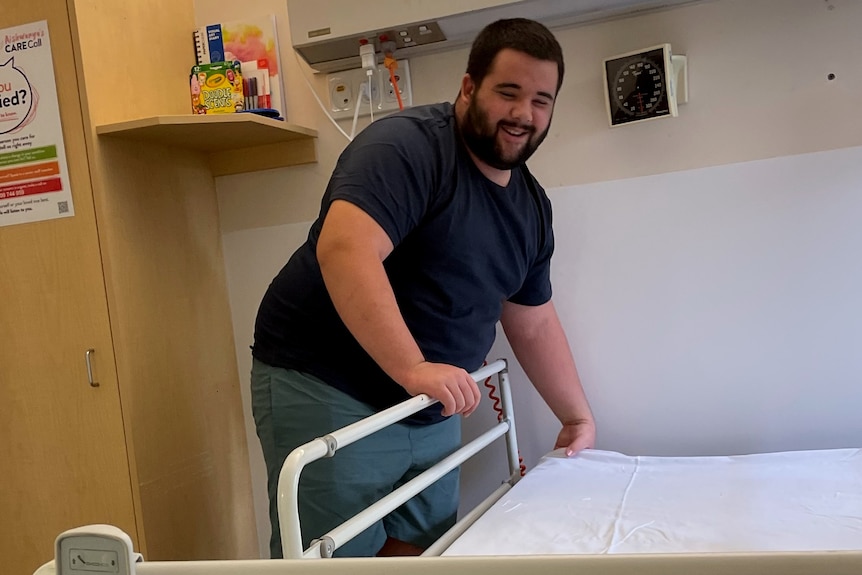 A young man, with black hair and a beard, is making his bed at a hospital. He's smiling.