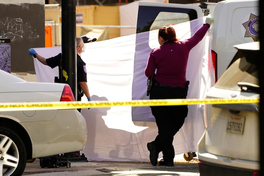 Two armed women, one a police officer and one a detective, hold up a white sheet across the boot of a white van.
