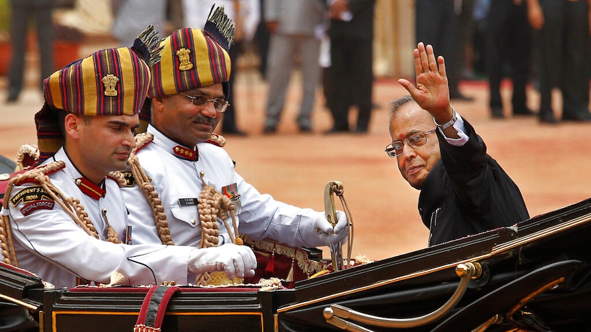 An elderly man is riding in a horse-drawn carriage next to two traditional Indian guards who look to the camera.