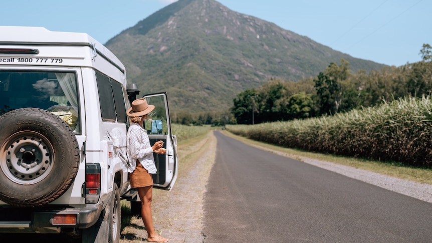 A woman stands next to a troop carrier near a cane field and big mountain.