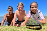 Three children, wet with river water, pose on the banks with a long-necked turtle with algae on its shell