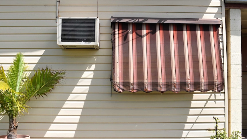 The side of a house with an air conditioning unit and an awning pulled down over the window on a sunny day.