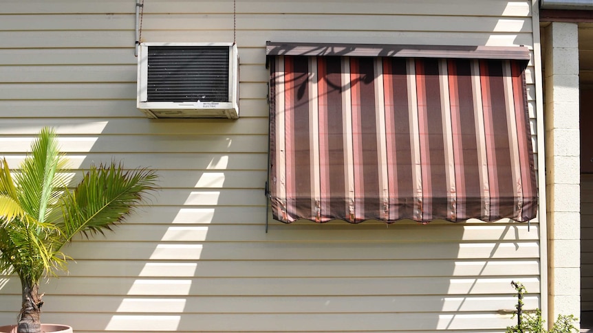 The side of a house with an air conditioning unit and an awning pulled down over the window on a sunny day.