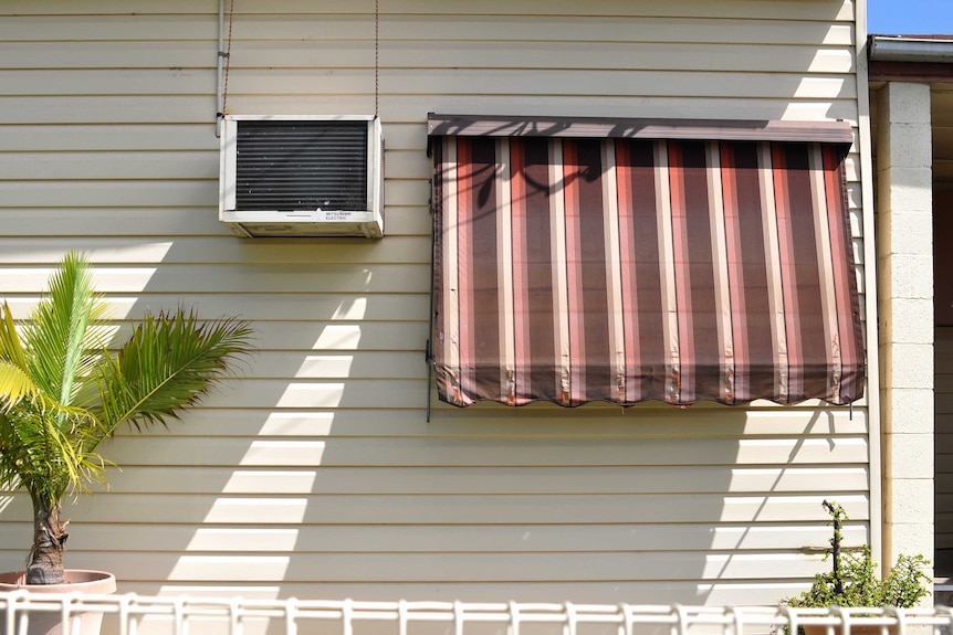 The side of a house with an air conditioning unit and an awning pulled down over the window on a sunny day.