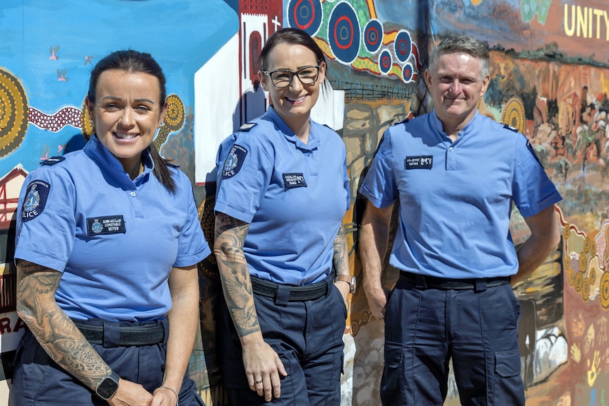 Two female and one male police officers stand against a colourful wall smiling, wearing blue uniform. 