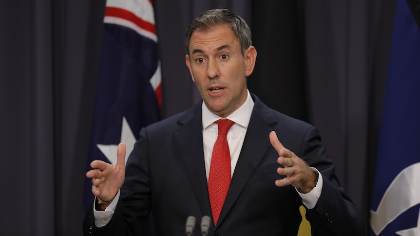 A medium shot of a man in a suit and tie, gesturing with his hands and speaking in front of the Australian flag.