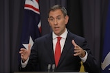 A medium shot of a man in a suit and tie, gesturing with his hands and speaking in front of the Australian flag.
