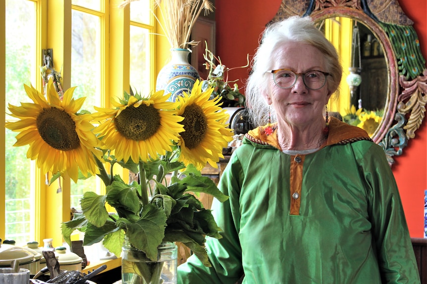 She wears a green silk robe and has white hair. She stands beside a vase of sunflowers and yellow window panes