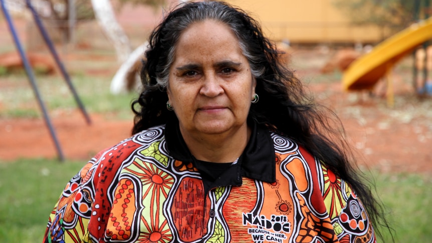 She looks at the camera, her dark curly hair is loose. She wears a colourful top with an Aboriginal art design. 