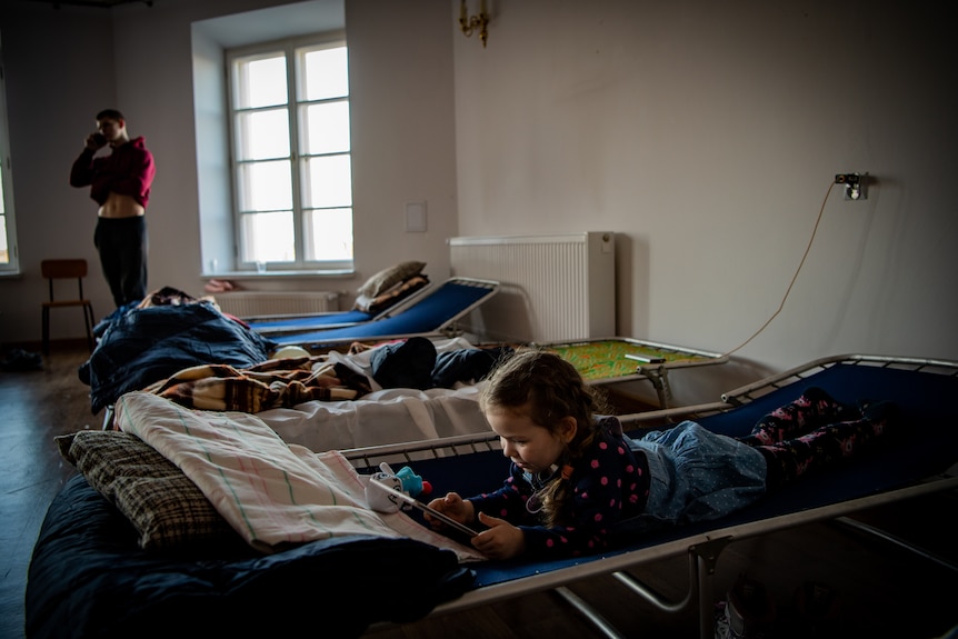 A girl lies on a makeshift bed looking at a device as her brother, in the background, speaks into a phone