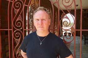A man in a black t-shirt wearing a cross around his neck leans against a church gate.