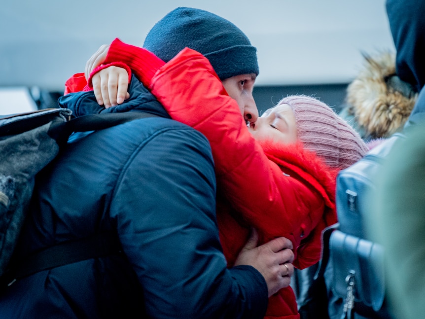 A little girl in a red coat and pink beanie wraps her arms around her Dad's neck, kissing his cheek
