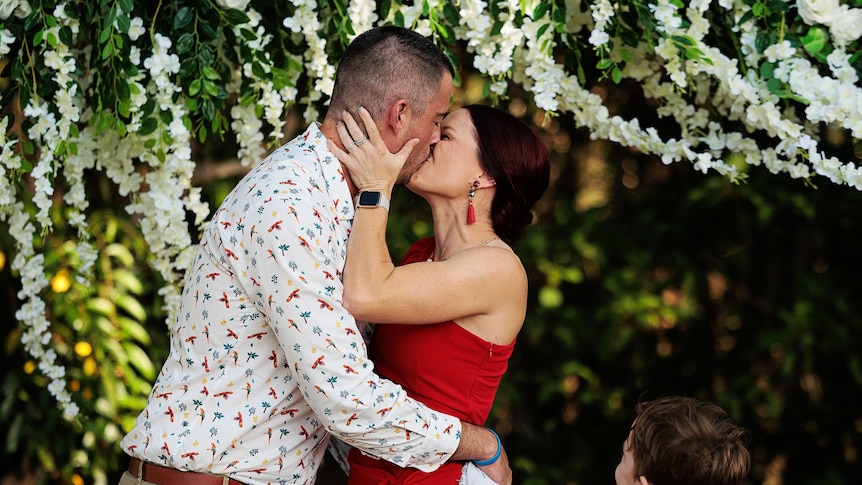 A woman in a red dress and a man kissing under a flowery arbor.