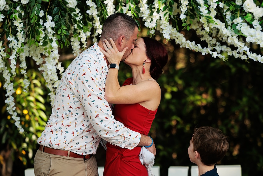 A woman in a red dress and a man kissing under a flowery arbor.