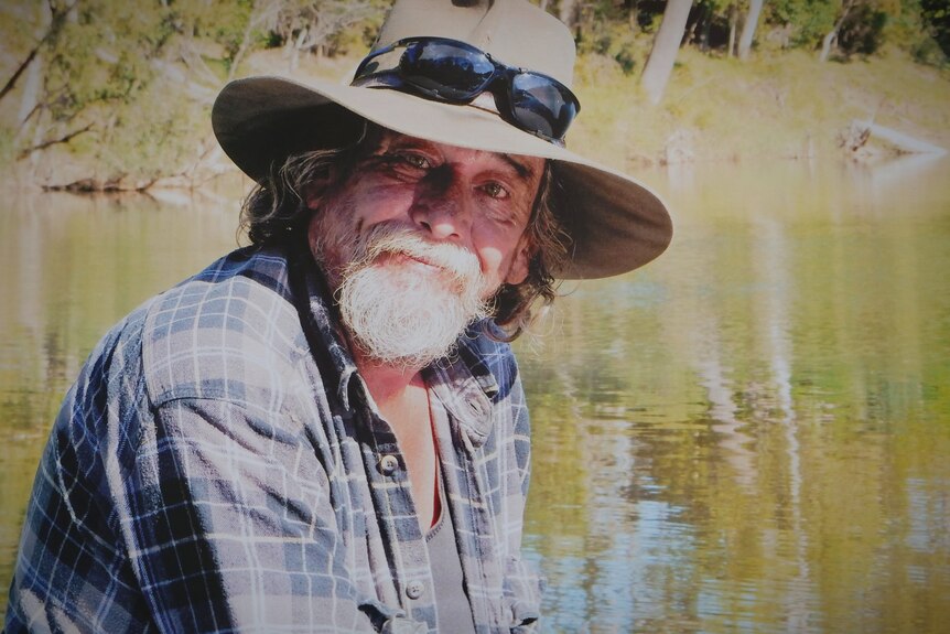 A bearded man in front of a river wearing a wide brim hat slightly smiling at the camera