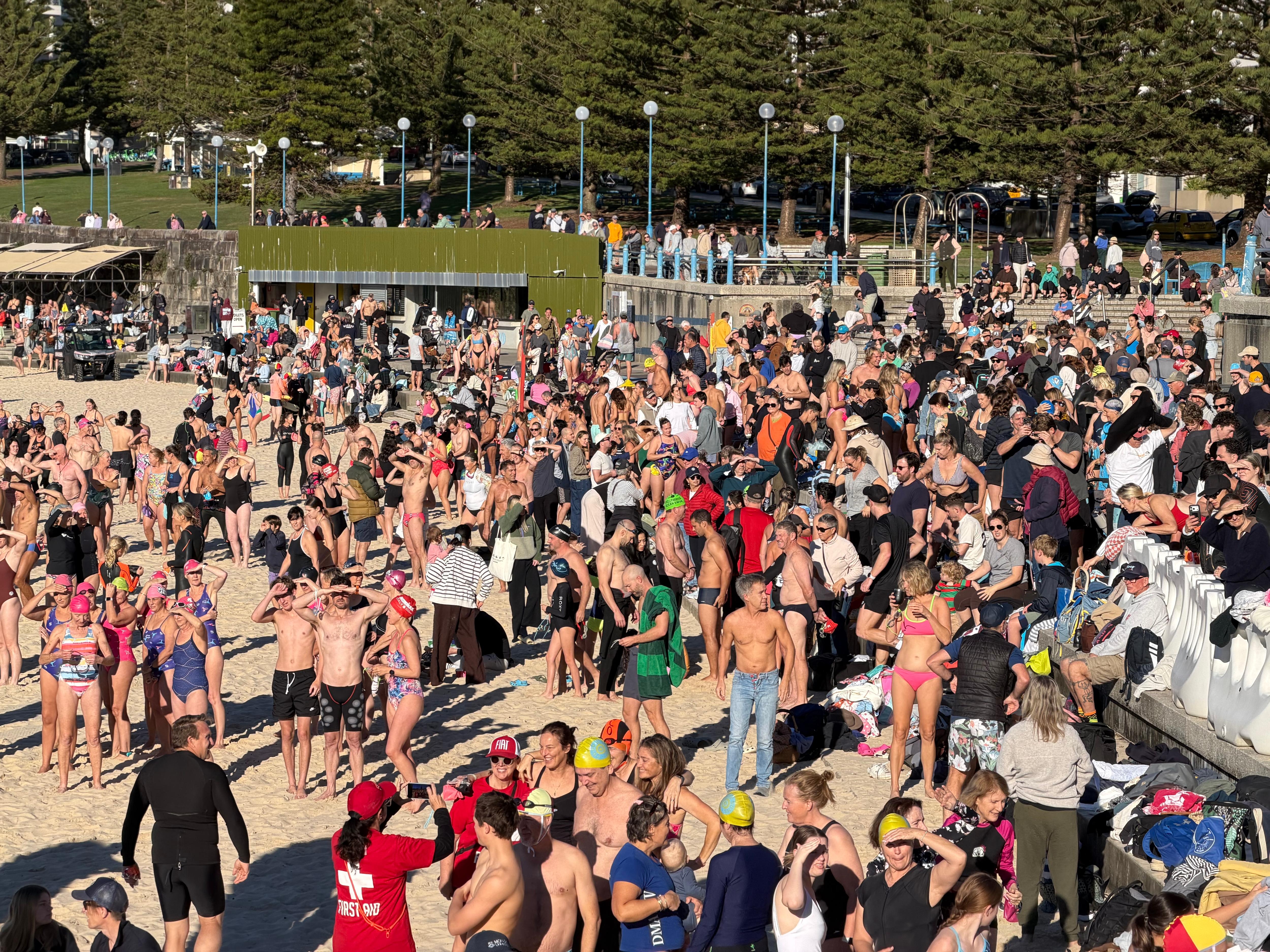 A large crowd of people in swimwear gathered on the sand and a pathway next to the beach on a sunny morning.