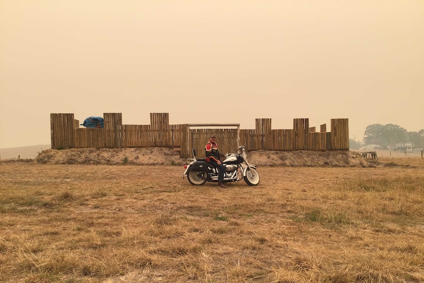 A man sits happily on a Harley Davidson motorbike in front of a homemade fort on a day shrouded in yellow haze.