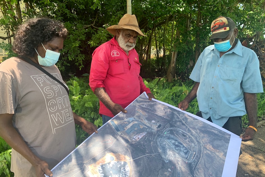 Three people look at a map of a mine and look serious. They are wearing face maks.