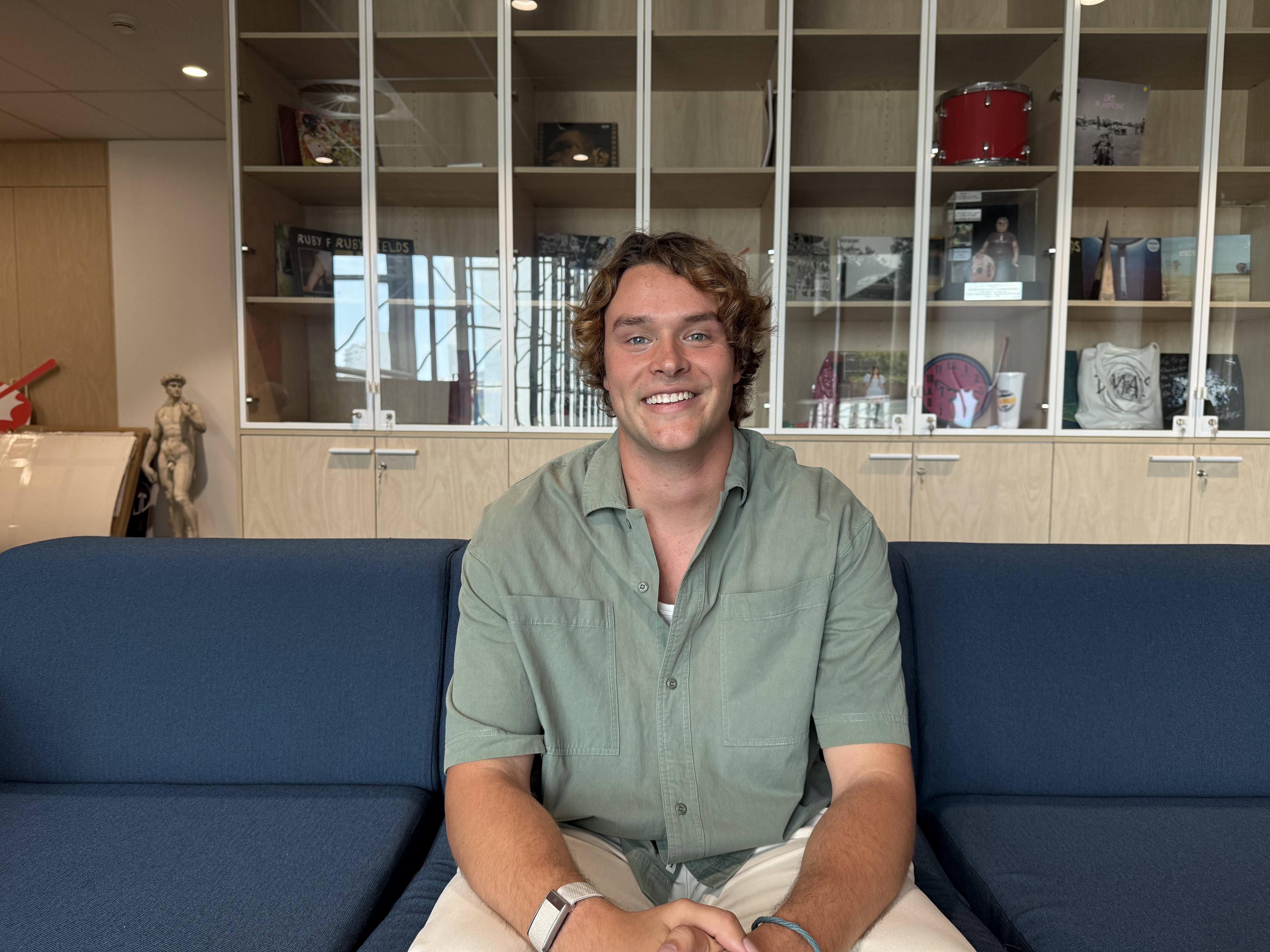 Um homem branco de cabelos loiros está sentado, sorrindo e olhando para a câmera, vestindo uma camisa verde de mangas curtas.