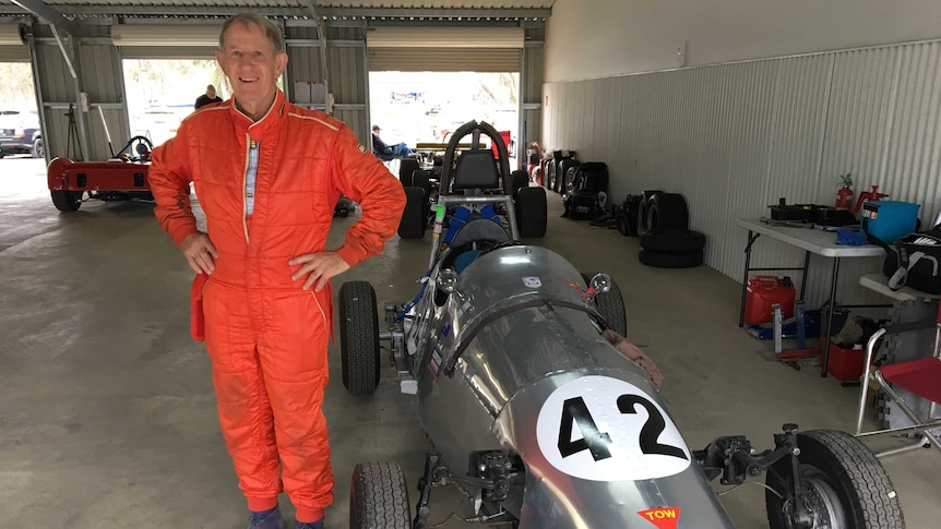 A man stands in an orange racing suit next to a Morris Special racing car.