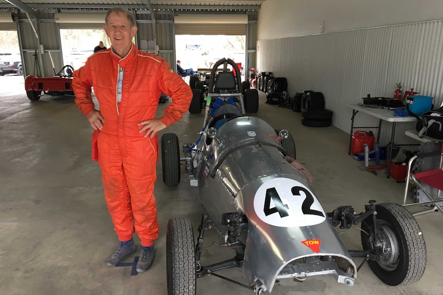 A man stands in an orange racing suit next to a Morris Special racing car.