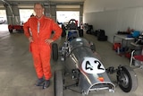 A man stands in an orange racing suit next to a Morris Special racing car.
