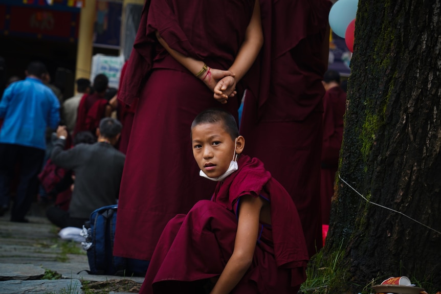 A boy in maroon robes, wearing a surgical mask on his chin, sits on the ground next to a tree
