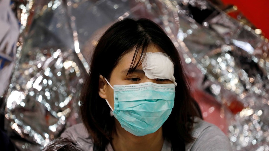 A young girl with a mask on her face and a dressing on her forehead sits wrapped in space blanket.