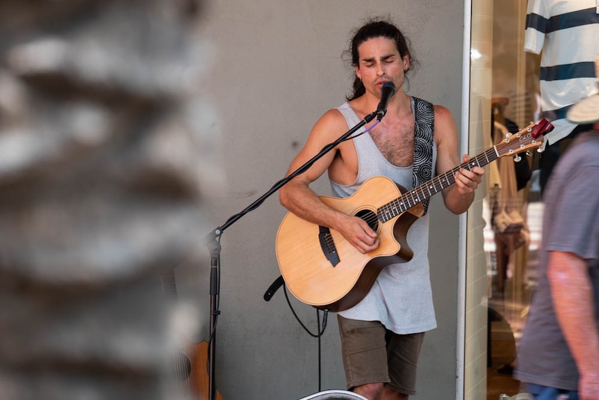 A busker playing guitar and singing into a microphone on the street in Tamworth.