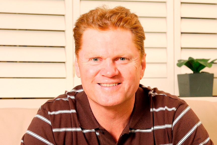 A man wearing a brown polo shirt with white horizontal lines smiles as he sits and faces the camera.