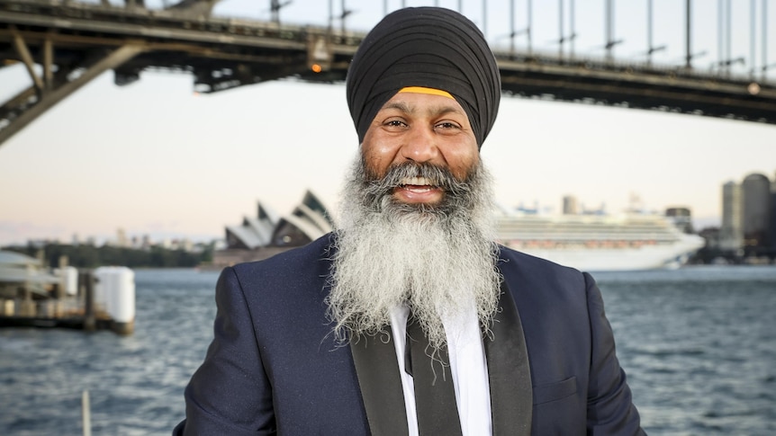 A man in a turban with a beard smiles with a trophy in front of the Sydney Harbour Bridge. 