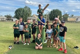 A bunch of kids, some barefoot, gather on a green cricket pitch while two are up on a big green bin with cricket bats in hand.