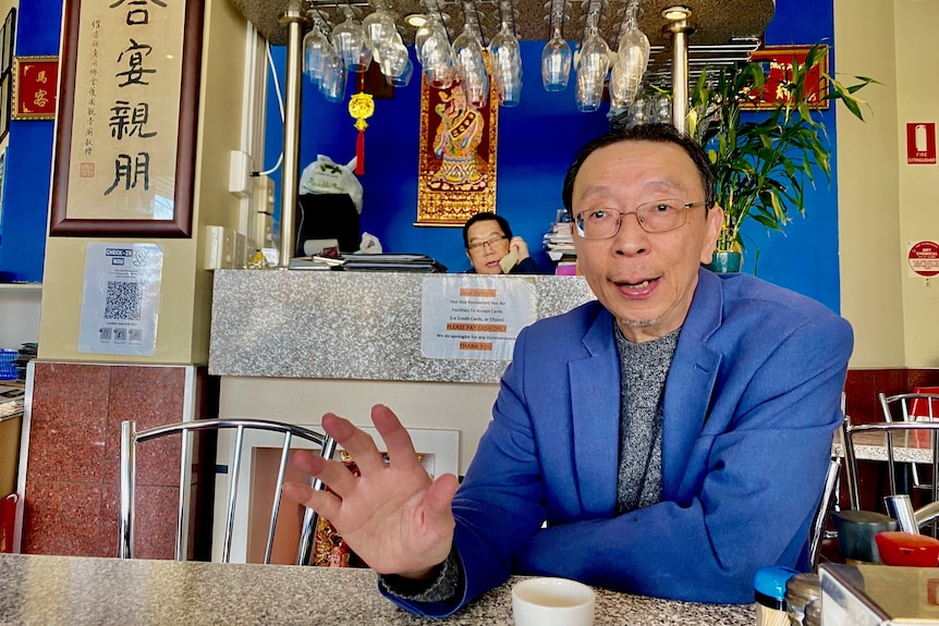 An older Vietnamese man sits in a restaurant at the table with the owner behind him