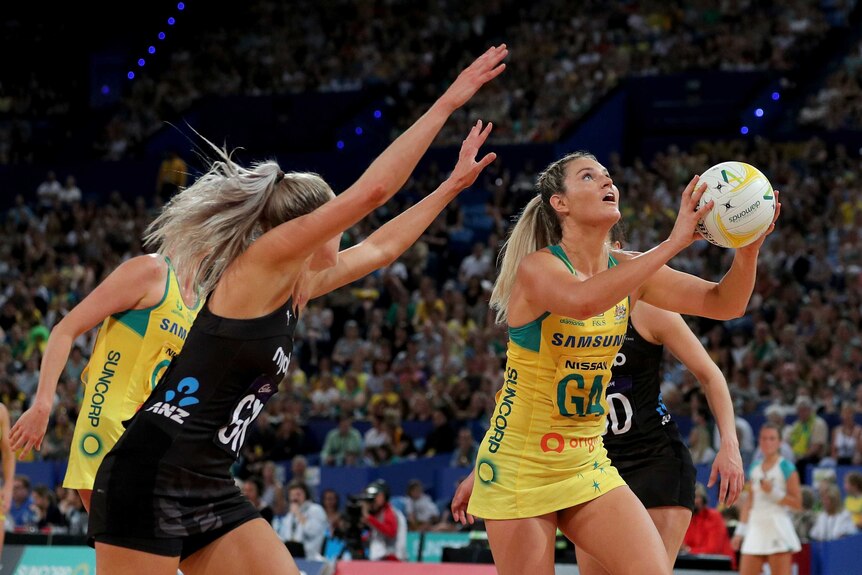 An Australian netballer looks up while shooting as a New Zealand defender stretches to block it.