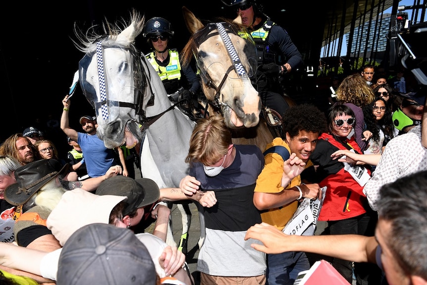 Two police officers on horses charge a crowd of protesters outside the Melbourne Exhibition Centre on a sunny day.