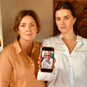Two women looking at the camera while holding a phone displaying a photo of an elderly woman.