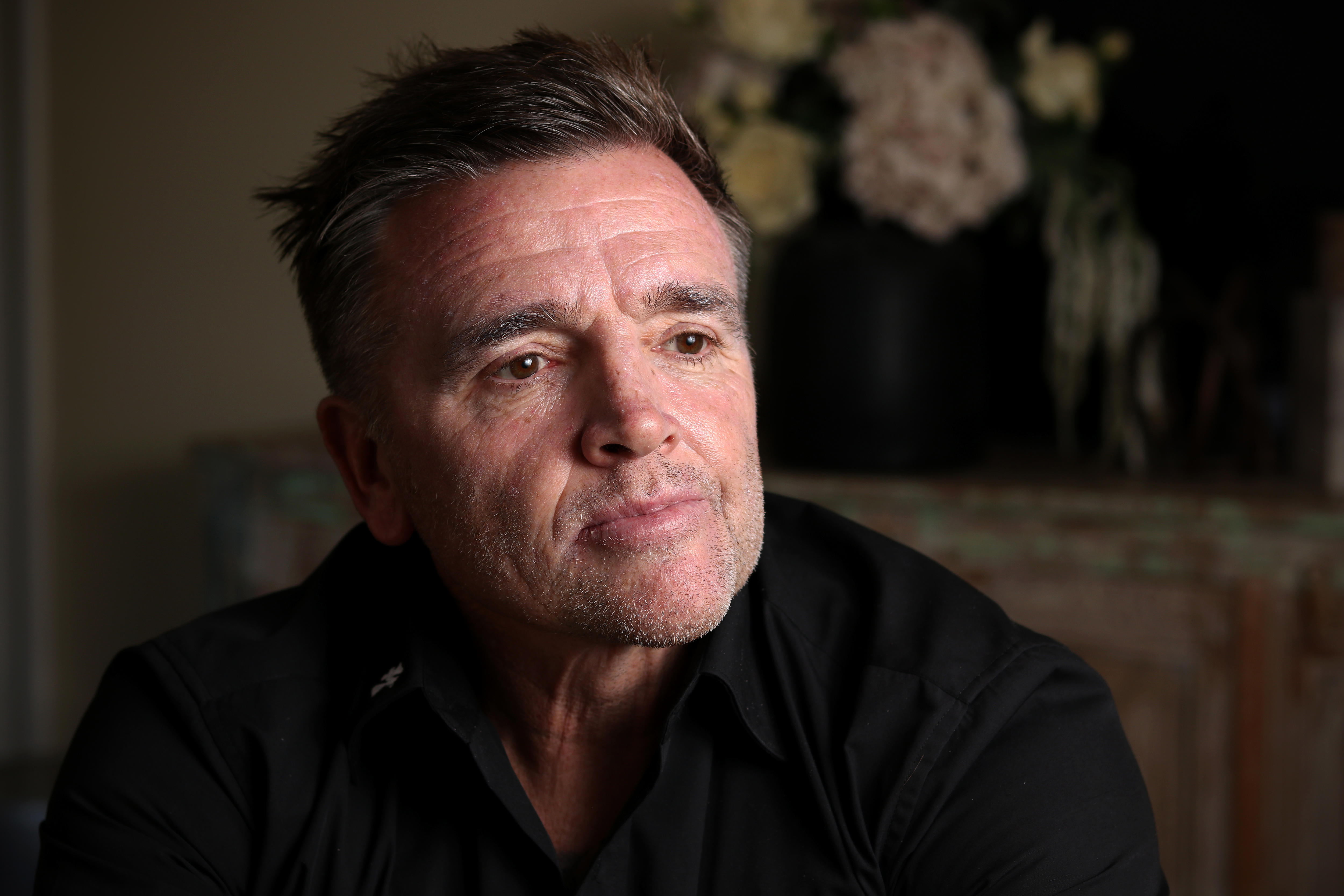 A close up of Jay Allan wearing a black shirt sitting looking into the distance while sitting in a living room 