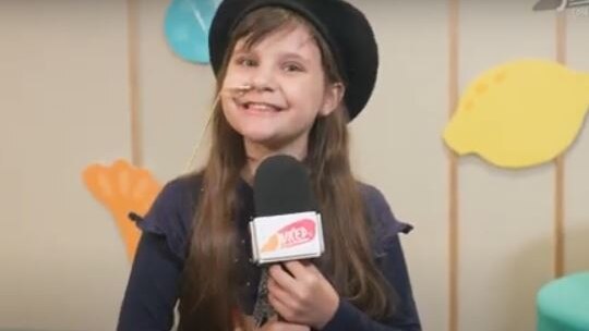 A young girl with a nasal tube wears a beret and smiles broadly as she holds a microphone in a studio.