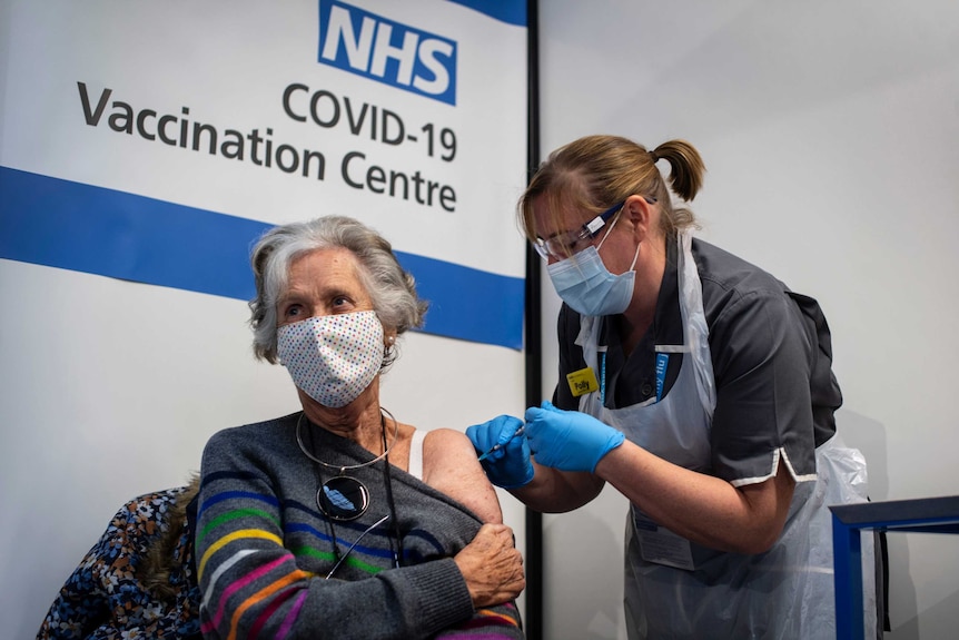 A woman gives a seated woman an injection in her shoulder.