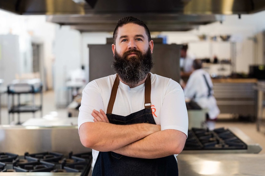 Man in apron stands with arms folded in commercial kitchen