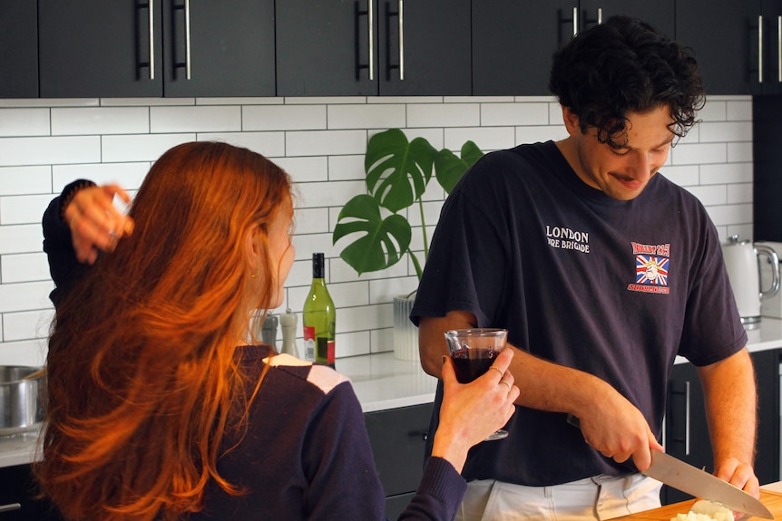 A woman with long red hair holds a glass of wine as she watches a man cut food in the kitchen. He's smiling.