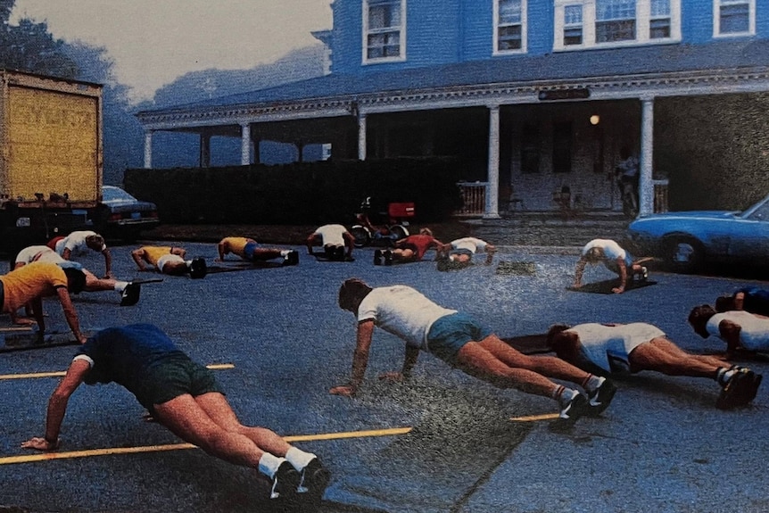 A grainy old photo of several men doing push ups outside a grand old house.