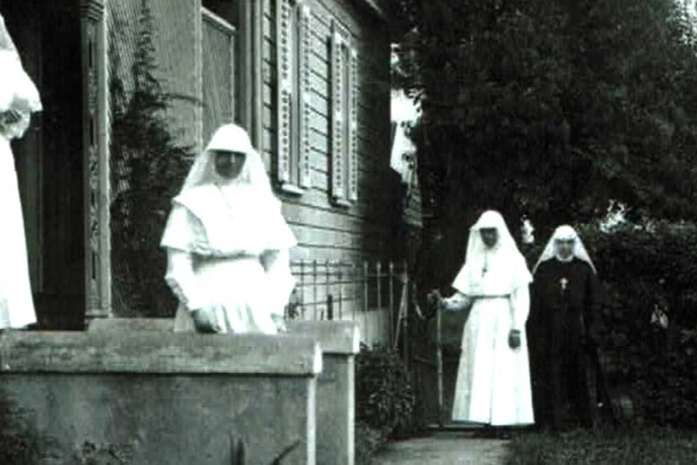 An old black and white photo of nuns wearing all white uniforms outside timber and stone building.