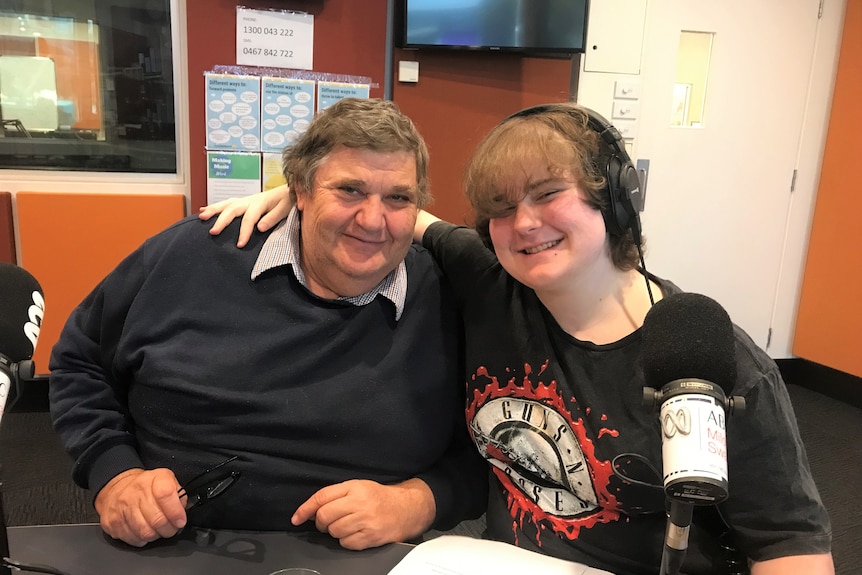 A man and his son smile at the camera. They both have light brown hair. They are sitting in a radio studio with a microphone.