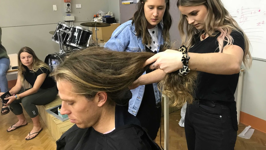 A teacher with long hair sits in a music room with hairdresser and student assessing hair.